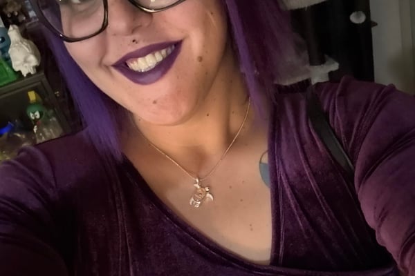 Person with purple hair and glasses smiling at camera indoors