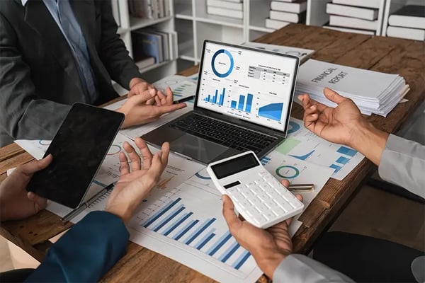 Business team reviewing financial data on laptop, tablet, and calculator at wooden desk with charts and documents