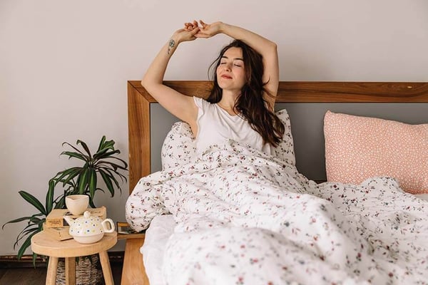 Woman stretching in bed with floral bedding, wooden headboard, and potted plant on nightstand