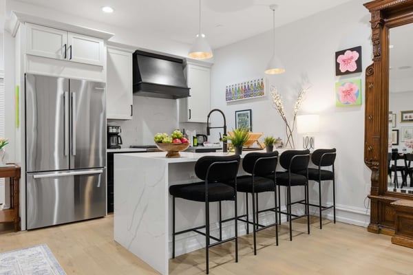 Modern kitchen with white cabinetry, stainless steel appliances, marble island with black bar stools, and pendant lighting